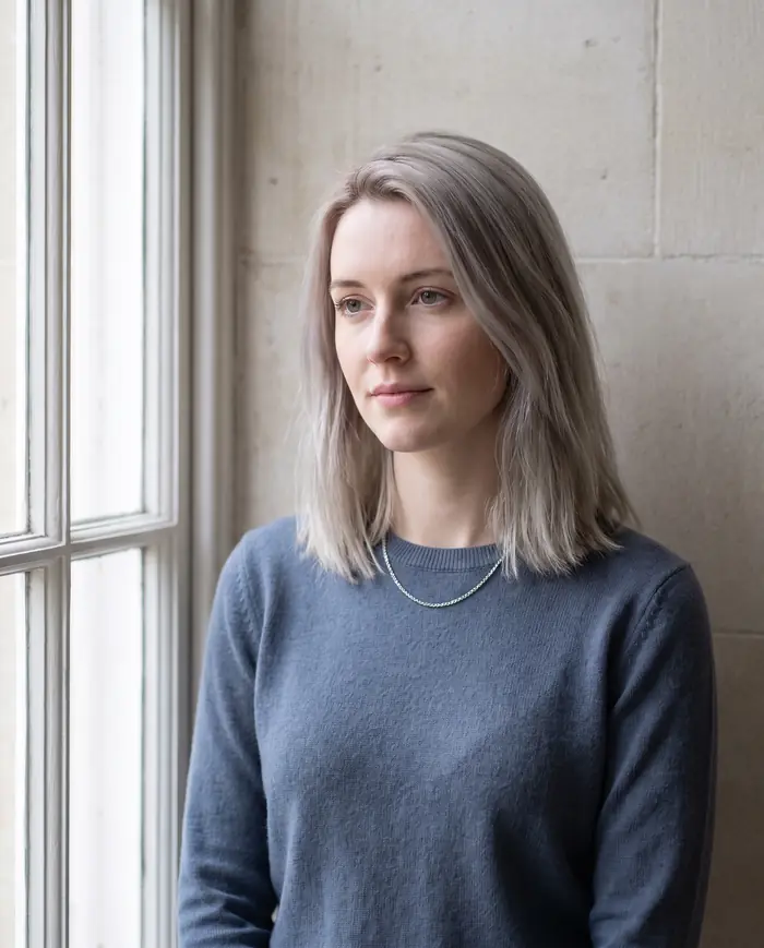 Woman with straight ash blonde hair in a slate blue sweater, standing by a window